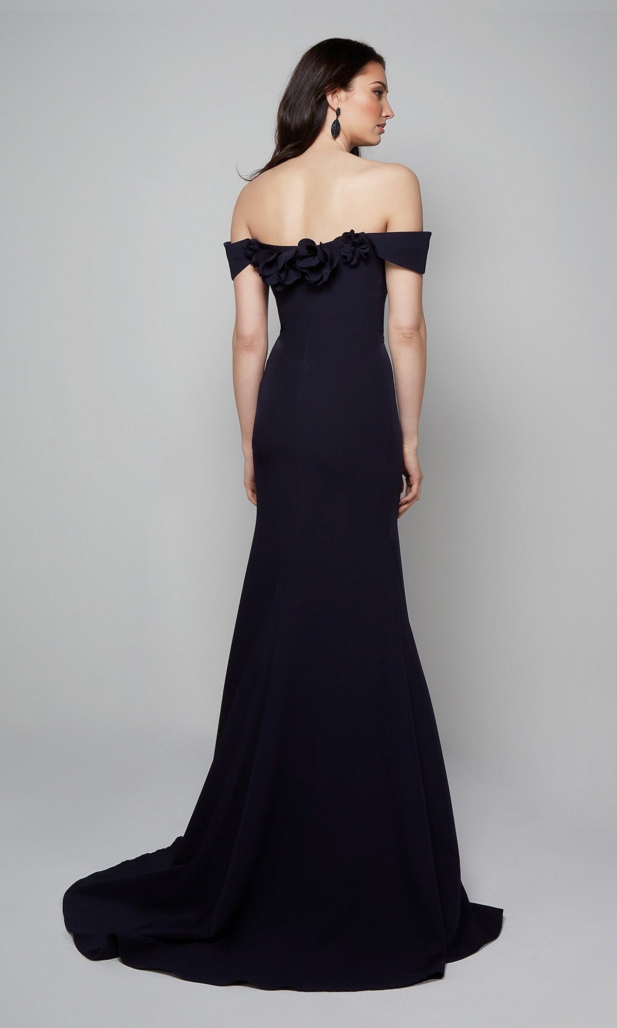 A woman wears the Alyce 7062 Long Formal Dress with an off-the-shoulder neckline and 3D floral back accents, posed against a plain light gray background.